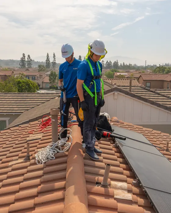 Two Titan crew members in helmets and harnesses on a clay tile roof next to solar panels